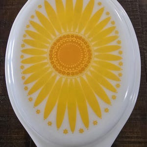 May include: White oval Pyrex lid featuring a yellow sunflower design. The sunflower has a bright orange centre and yellow petals. Small yellow flower accents surround the edge. A vintage kitchenware item.