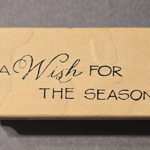 Puede incluir: Un sello rectangular de madera con las palabras "A Wish FOR THE SEASON" en tinta negra. La palabra "Wish" está en una fuente de escritura decorativa. El sello es de color marrón claro y está sobre una superficie gris.
