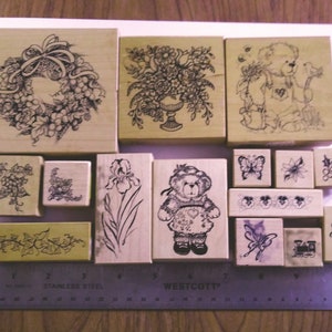 May include: A collection of 14 wooden rubber stamps with various designs, including a wreath, flowers, a teddy bear, butterflies, and a train. The stamps are arranged on a wooden surface with a ruler.