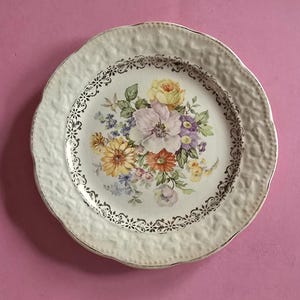May include: A vintage white ceramic plate with a textured rim and gold trim. The centre features a floral bouquet in shades of yellow, purple, orange, and pink. The plate has a scalloped edge and a decorative border.
