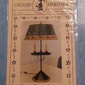 May include: A Country Heritage lamp design, No. 201, featuring a black lamp with a green lampshade adorned with a fruit and leaf pattern. The design includes two candle-style lights and is designed by Helen Jeglic.