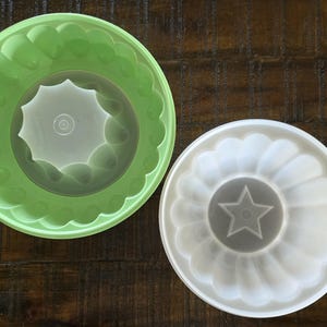 May include: Two vintage plastic jelly moulds. One is light green, the other is white. Both have a fluted design around the edges and a decorative centre. The green mould has a star-shaped centre, while the white mould has a star-shaped centre.