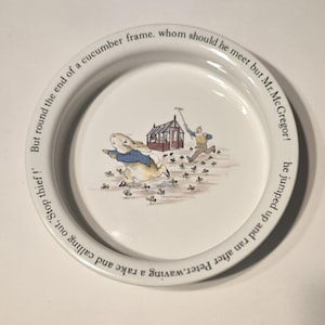 Puede incluir: Plato de cerámica blanca con borde de texto, que representa una escena de un cuento infantil. La ilustración central muestra un conejo huyendo de un hombre con un rastrillo cerca de un pequeño edificio.