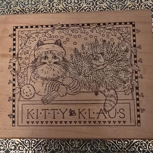 May include: A wooden stamp featuring a cat wearing a Santa hat and holding a log, with a Christmas tree. The text "KITTY KLAUS" is displayed below the cat. The design is surrounded by holly and berries.