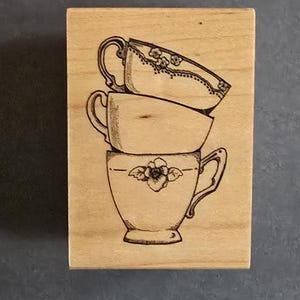 May include: A wooden stamp featuring a stack of three teacups. The teacups are illustrated in black ink, with floral details on the top and bottom cups. The stamp is rectangular and made of light-colored wood.