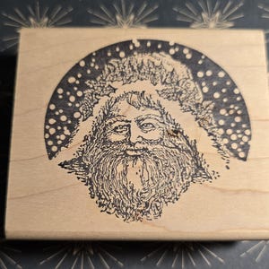 May include: A rectangular wooden stamp featuring a detailed illustration of Santa Claus. The black and white image shows Santa's face, beard, and a snowy background. The stamp is likely used for crafting or holiday-themed projects.