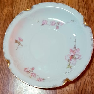 Może przedstawiać: Biała porcelanowa miska do serwowania z falistym brzegiem i złotym wykończeniem. Miska posiada delikatne różowe wzory kwiatowe. Element vintage do serwowania lub ekspozycji, na drewnianej powierzchni.