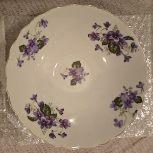 Puede incluir: Un platillo de cerámica blanca con un borde dorado y un diseño floral con violetas moradas.