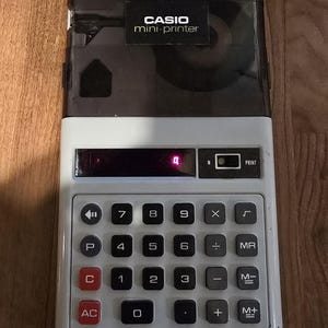Pode incluir: Uma calculadora mini-impressora Casio vintage, branca com a parte superior preta. Possui um teclado numérico e botões de função. O visor mostra o número 4. A mini-impressora Casio está conectada a uma fonte de alimentação.