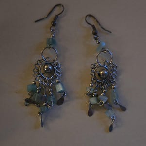 May include: A pair of silver-toned dangle earrings. Each earring features a decorative filigree design with a central silver bead, small blue-green beads, and dangling teardrop-shaped charms. The earrings have a hook closure.