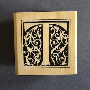 May include: A wooden stamp featuring the letter "T" in a black, ornate, vintage design. The letter is framed within a rectangular border. The stamp has a light brown color.