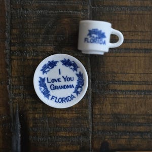 May include: A white ceramic miniature teacup and saucer set. The saucer reads "I Love You Grandma FLORIDA" in blue script. The teacup has "FLORIDA" in blue. The set is on a dark wood surface.