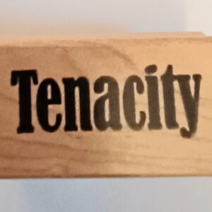 May include: A wooden stamp with the word "Tenacity" printed in black ink.