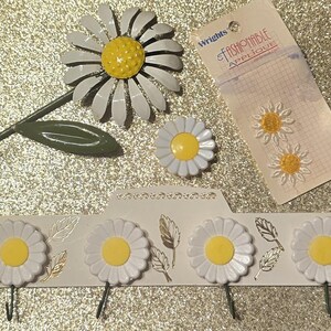 May include: A metal daisy wall hook with a green stem, a single daisy-shaped button, a package of two Wrights Fashionable Applique daisy patches, and a white wall-mounted rack featuring four daisy-shaped hooks with gold leaf accents.