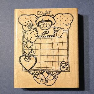 May include: A wooden stamp featuring a whimsical angel design. The angel has dotted wings, a bow, and holds a grid-patterned sign. A heart and button embellishments add detail to the black and white illustration.