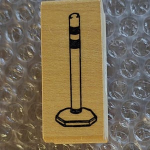 May include: A rectangular wooden stamp featuring a black line drawing of a bollard. The bollard has a round top, a striped section, a long stem, and a hexagonal base. The stamp is on a bubble wrap surface.
