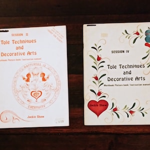 Könnte beinhalten: Zwei weiße Lehrbücher mit orangefarbenen Rändern mit dem Titel "Tole Techniques and Decorative Arts". Das Buch links zeigt eine rot-weiße Illustration eines Herzens und zweier Vögel. Das Buch rechts zeigt ein rotes Herz mit grünen und goldenen floralen Designs und einem blauen Vogel.