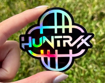 May include: A black holographic sticker with the word "HUNTR/X" in white, rainbow-colored lettering. The sticker has a unique shape with a series of vertical and horizontal lines. The background is blurred green grass.