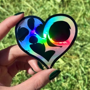 May include: A heart-shaped holographic sticker with a black paw print design. The sticker has a rainbow sheen and is held against a green background. The sticker's edges are black.