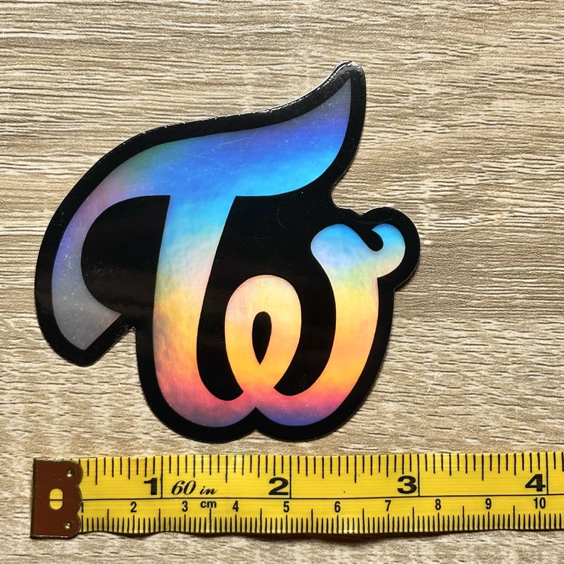 May include: A holographic sticker with the word "TWICE" in a stylized font. The sticker is black with a rainbow iridescent finish.