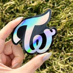 May include: A holographic sticker with the word "Twice" in a stylized font. The sticker is black with iridescent rainbow colors.