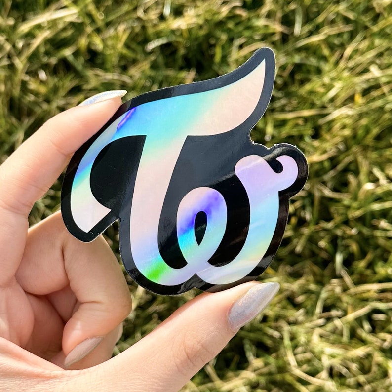 May include: A holographic sticker with the word "Twice" in a stylized font. The sticker is black with iridescent rainbow colors.