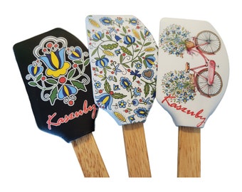 Può includere: Tre spatole con manico in legno e teste in silicone decorative. Una spatola ha una testa nera con un motivo floreale e la scritta "Kaszuby". Le altre due spatole hanno teste bianche con motivi floreali e di biciclette, anch'esse con la scritta "Kaszuby".