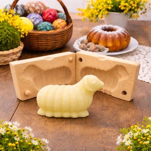 May include: A light yellow sheep-shaped butter sculpture sits in front of a wooden mold. The mold is in two pieces, with the sheep's form carved into each. A basket of decorated Easter eggs and a cake are in the background.