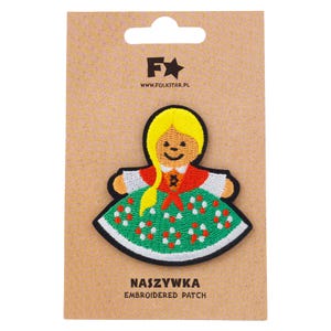 May include: Embroidered patch featuring a smiling girl in traditional Polish folk attire. The patch has a yellow hairstyle, a red top, and a green skirt with white and red floral accents. The patch is on a brown card with the text "NASZYWKA EMBROIDERED PATCH".