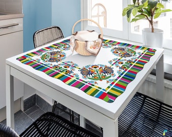 May include: A white tablecloth with a colorful folk art design featuring roosters and floral patterns. The border has a wavy edge with a striped pattern in various colors. A wicker basket sits on the table.