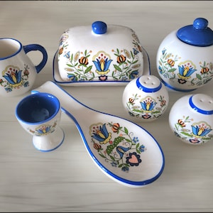May include: A white ceramic kitchen set with blue trim and floral designs. The set includes a creamer, butter dish, sugar bowl, salt and pepper shakers, an egg cup, and a spoon rest. The floral pattern features blue, yellow, and orange flowers.