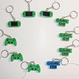 May include: A set of 8 green and blue keychains with different video game themed designs. The keychains feature a handheld game console, a game controller, and the words "Game On".