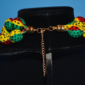Puede incluir: Un collar de m&uacute;ltiples hilos hecho de bolas de tela en verde, amarillo y rojo con un cierre de cadena dorada.