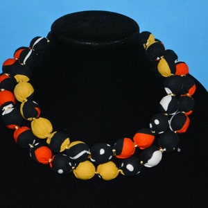 Puede incluir: Un collar de doble cadena hecho de cuentas de tela. Las cuentas son negras, blancas, naranjas y amarillas con patrones geom&eacute;tricos. El collar se muestra en un maniqu&iacute; negro.