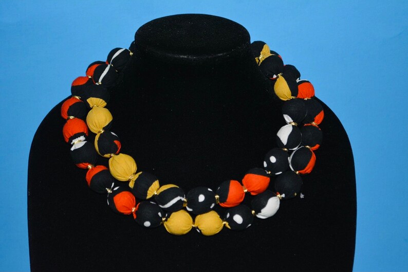 Puede incluir: Un collar de doble cadena hecho de cuentas de tela. Las cuentas son negras, blancas, naranjas y amarillas con patrones geom&eacute;tricos. El collar se muestra en un maniqu&iacute; negro.