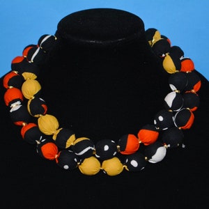 Puede incluir: Un collar de m&uacute;ltiples hilos hecho de cuentas cubiertas de tela. Las cuentas son en tonos de negro, blanco, naranja y amarillo. El collar est&aacute; en un maniqu&iacute; negro.