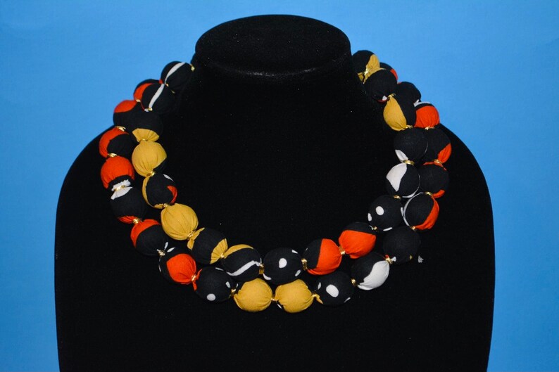 Puede incluir: Un collar de m&uacute;ltiples hilos hecho de cuentas cubiertas de tela. Las cuentas son en tonos de negro, blanco, naranja y amarillo. El collar est&aacute; en un maniqu&iacute; negro.