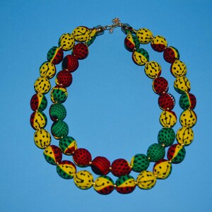 Puede incluir: Un collar de doble cadena hecho de cuentas cubiertas de tela. Las cuentas son amarillas, verdes y rojas con lunares negros. El collar tiene un cierre dorado.