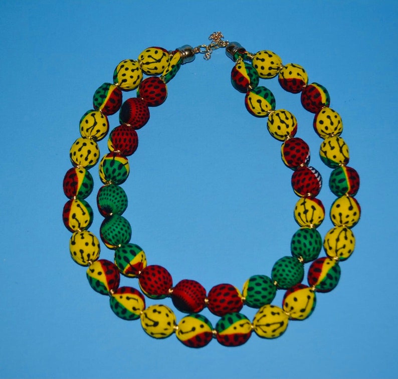 Puede incluir: Un collar de doble cadena hecho de cuentas cubiertas de tela. Las cuentas son amarillas, verdes y rojas con lunares negros. El collar tiene un cierre dorado.