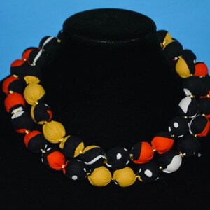 Puede incluir: Un collar de doble cadena hecho de cuentas de tela. Las cuentas son negras, blancas, rojas y amarillas con patrones geom&eacute;tricos.