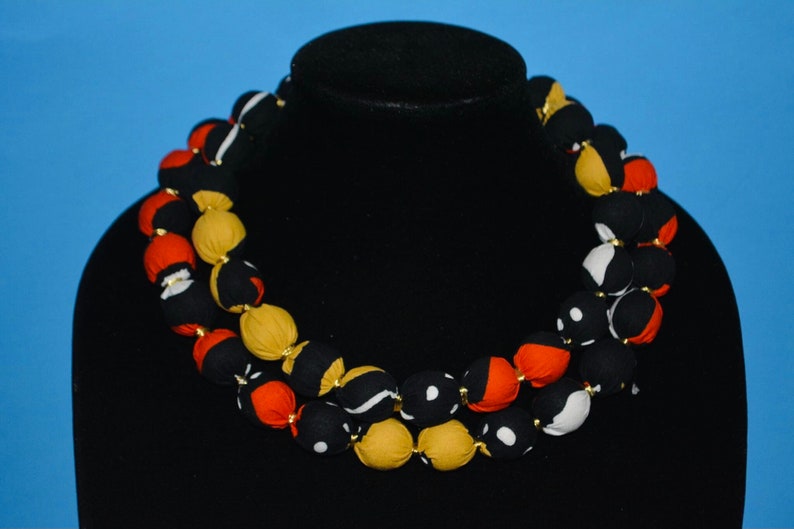 Puede incluir: Un collar de doble cadena hecho de cuentas de tela. Las cuentas son negras, blancas, rojas y amarillas con patrones geom&eacute;tricos.