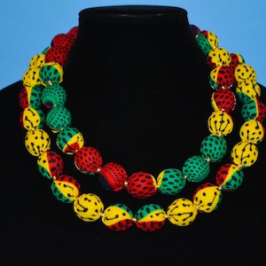 Puede incluir: Un collar de doble cadena hecho de cuentas grandes y redondas. Las cuentas est&aacute;n cubiertas de tela con un patr&oacute;n amarillo, verde y rojo. El collar se muestra en un maniqu&iacute; negro.