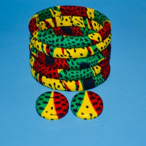 Puede incluir: Un conjunto de cinco pulseras de tela coloridas con un patrón verde, amarillo y rojo. Las pulseras están apiladas una encima de la otra. Se incluyen dos pendientes de tela a juego con el mismo patrón.