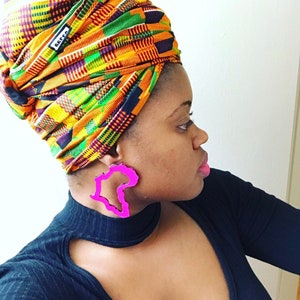 Puede incluir: Una persona con un turbante colorido y una camisa negra con grandes pendientes rosas con forma del continente africano.