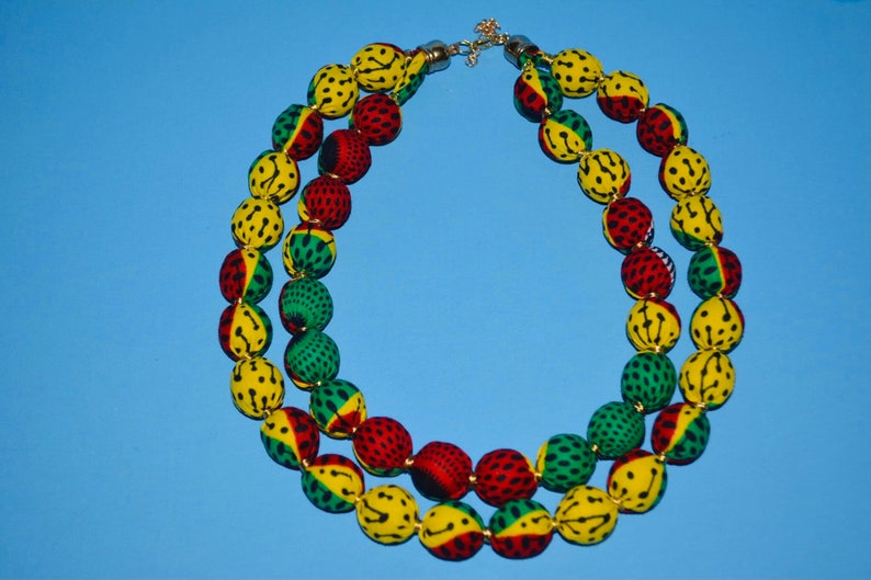 Puede incluir: Un collar de doble cadena hecho de cuentas de tela. Las cuentas son amarillas, verdes y rojas con un patr&oacute;n negro. El collar tiene un cierre dorado.