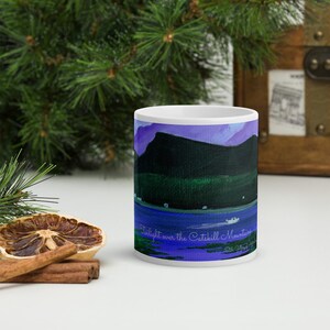 May include: White ceramic mug featuring a landscape painting of the Catskill Mountains at twilight. The artwork depicts a dark mountain range, a blue lake, and a purple sky. The mug has the text "Twilight over the Catskill Mountains".