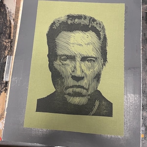 Puede incluir: Una impresión rectangular sobre tela verde oliva con un retrato en tinta negra de una persona con el pelo corto. La imagen está creada con líneas horizontales distintivas que definen los rasgos faciales, las sombras y los contornos de la cabeza.
