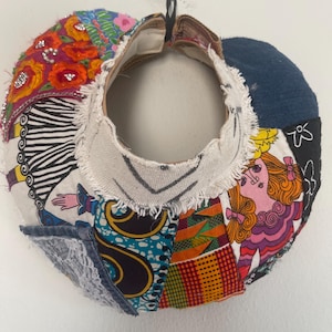 May include: A circular fabric collar featuring a patchwork of diverse patterns including floral prints, geometric shapes, and stylized figures. The piece incorporates denim, lace, and embroidered accents with frayed edges around the inner neckline.