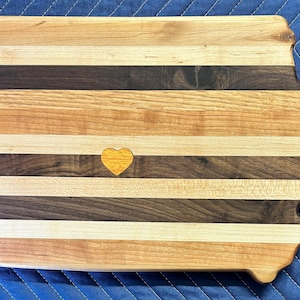 May include: A wooden cutting board shaped like the state of Iowa with a yellow heart in the center. The board is made of different types of wood, creating a striped pattern.