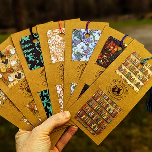 May include: A collection of eight colorful bookmarks with different patterns, including floral, geometric, and book-themed designs. Each bookmark is attached to a brown cardstock backing with a tassel. The bookmarks are labeled "Low Rollin' Art Creations".